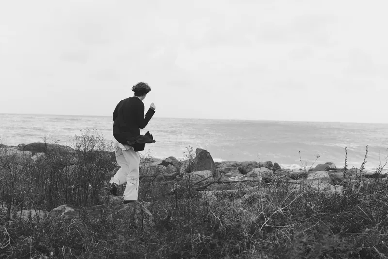Man walking silhouette along a rocky coastal shoreline in grayscale, capturing a quiet moment by the sea with a windswept atmosphere and rugged terrain under an overcast sky.