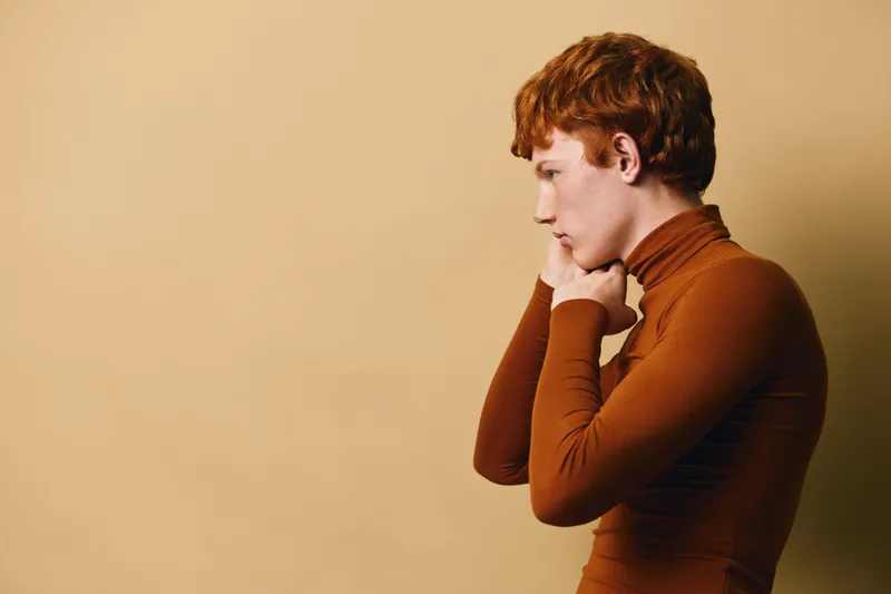 Profile of young redhead woman in brown turtleneck shirt with thoughtful expression against beige background in studio with soft lighting.