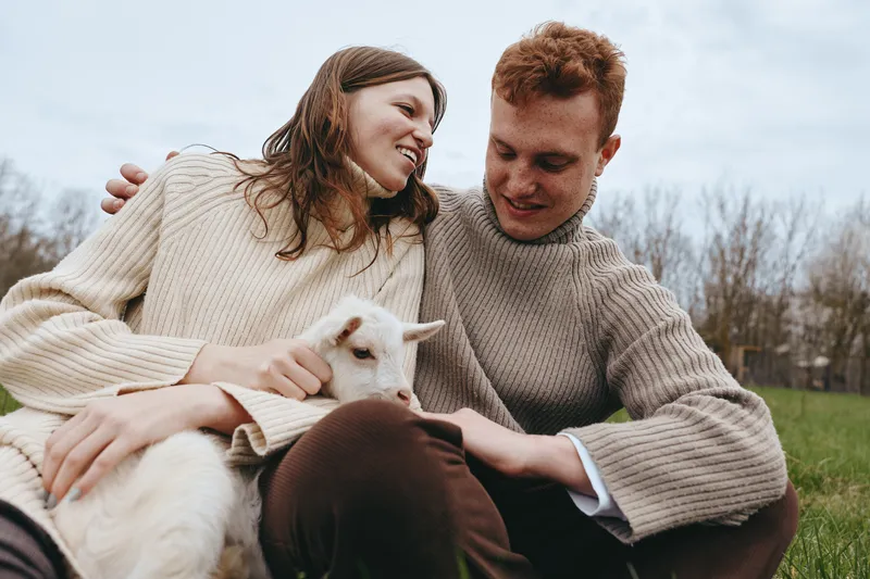 Couple outdoors reading with a goat kid, smiling and cozy in sweaters, a warm lifestyle moment amid meadow, grass and sunlight, sharing joy, calm connection, natural colors