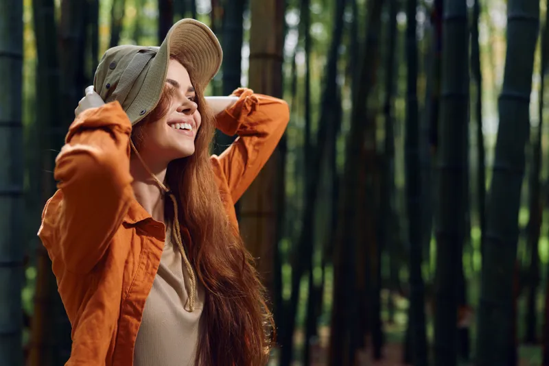 Woman smile hat hair bamboo forest portrait of a joyful young female with long redhead hair wearing a hat and jacket in sunlight among green bamboo grove outdoor lifestyle.