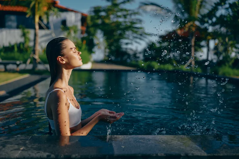 Woman relaxes in a sunny pool as water sprays from her hands, in a tropical outdoor setting, a calm lifestyle moment with bright light, casual swimwear, and serene summer mood.