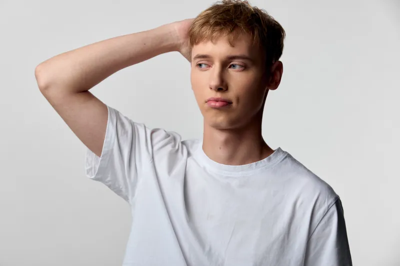 Thinking man in a casual white tee scratching his head, conveying curiosity, doubt and introspection in a clean studio setting with neutral backdrop and soft light