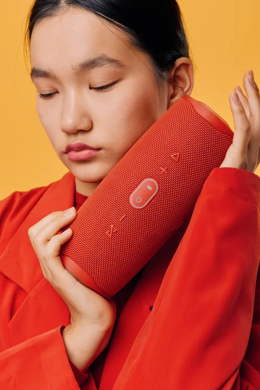 Woman holding red wireless speaker close to face with eyes closed wearing matching red jacket. Portrait of relaxed girl enjoying music with orange background and fabric texture on speaker.