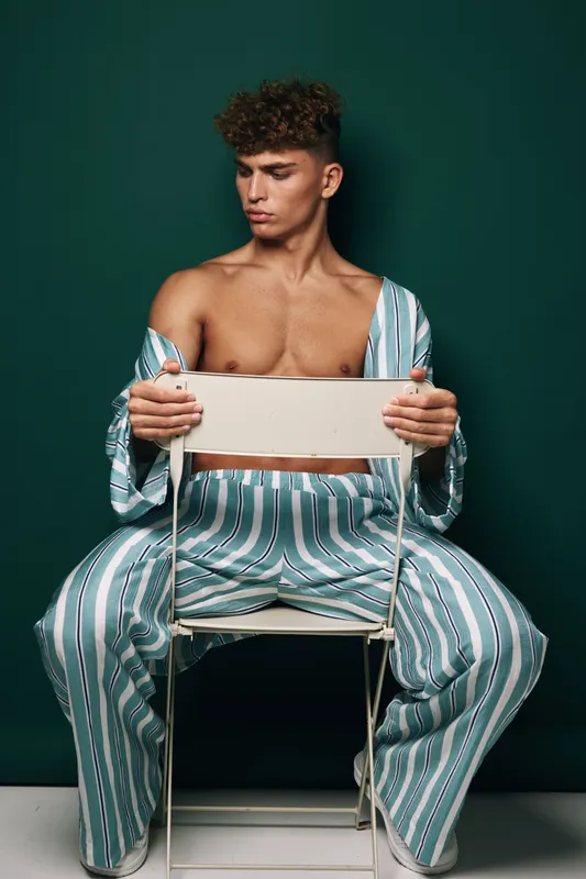 Young man with curly hair wearing striped pajamas sitting on white chair against dark green background, looking down with serious expression and relaxed posture.