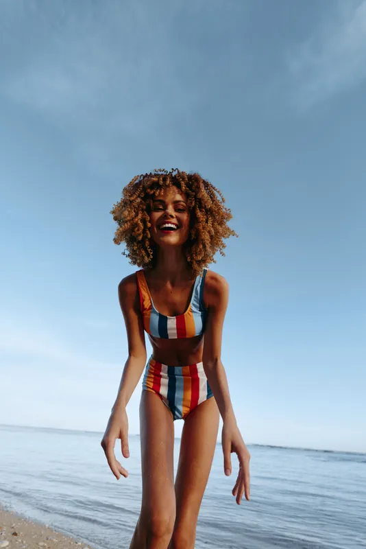 Woman with curly hair wears colorful striped swimwear on a quiet beach, ocean waves and blue sky behind, sunlit summer relaxation and freedom, joyful healthy lifestyle outdoors moment