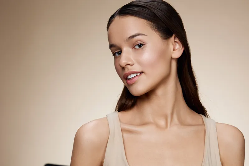 Beautiful young woman with long hair, natural makeup, and a confident smile, wearing a light colored top against a soft beige background, exuding calm and elegance