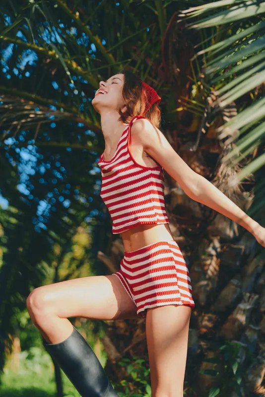 A carefree woman wearing a striped two piece outfit dances outdoors under sunny skies, arms extended, enjoying a lively summer moment in a lush garden setting.