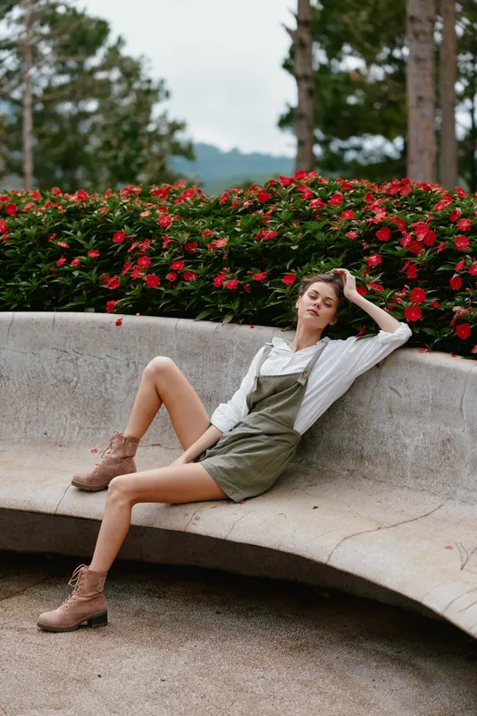 Stylish young woman in olive green outfit sitting on a bench surrounded by vibrant red flowers, expressing a relaxed mood in a natural setting