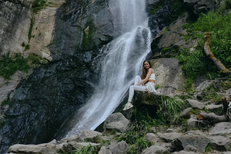 Young woman sitting on a rock near a cascading waterfall surrounded by lush green vegetation and natural rocky landscape in an outdoor adventurous setting.