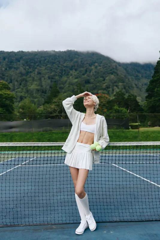 Young female tennis player in white athletic wear, smiling and posing on a tennis court with mountains in the background, embodying a dynamic and healthy lifestyle