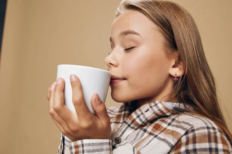 Girl with closed eyes holding white cup near face enjoying aroma of hot drink at cozy studio. Child in plaid shirt feeling relaxed with warm beverage closeup portrait.