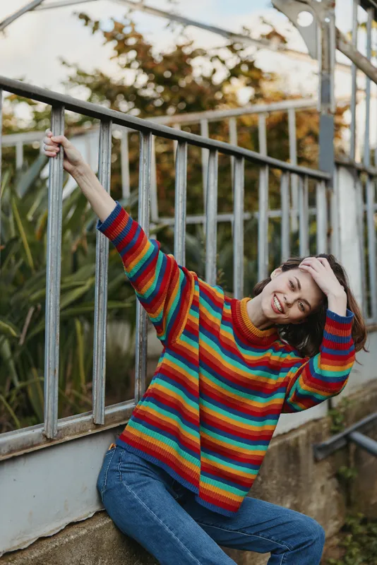 Woman in colorful striped sweater smiling in a casual portrait outdoors against a metal railing, joyful young model seated by greenhouse plants, fashion knitwear and relaxed mood.