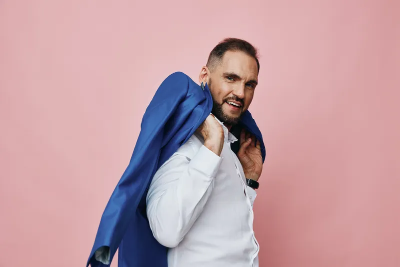 A confident business professional stands with a blue jacket draped over his shoulder, against a solid pink background, radiating focus, optimism, and poised energy.