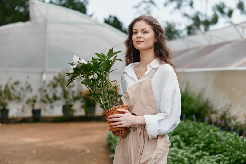 Young woman in a garden holding a pot of flowers, wearing a light shirt and aprons, conveying a joyful and serene vibe amidst greenery and greenhouses