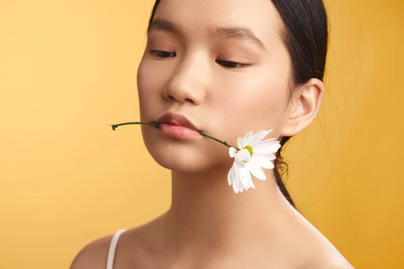 Asian woman holds a delicate flower between her lips against a soft yellow backdrop, creating a serene, minimalist portrait with calm mood and graceful poise.