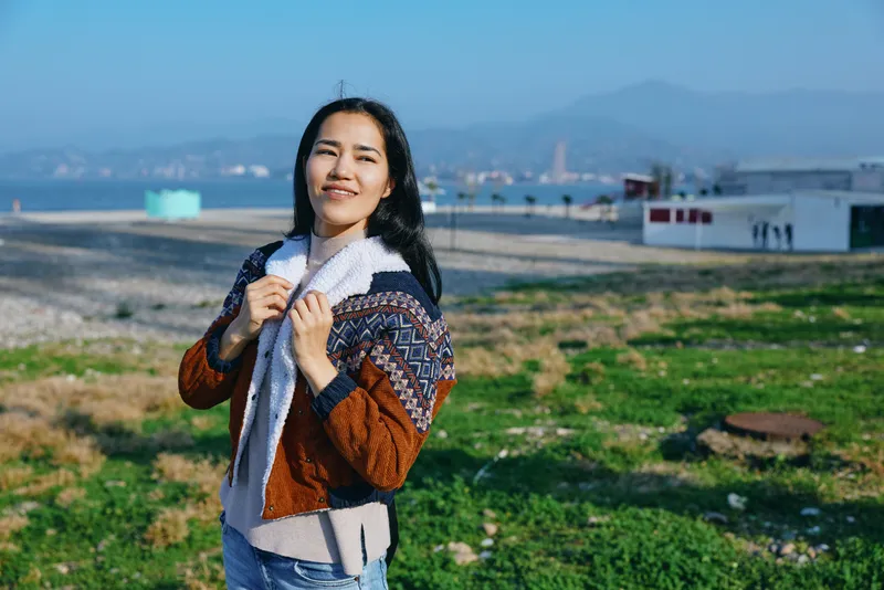 Woman wearing a cozy scarf and jacket stands outdoors in a grassy field near the coast, enjoying fresh air, sunlight and a relaxed, cheerful mood with distant hills and blue sky.