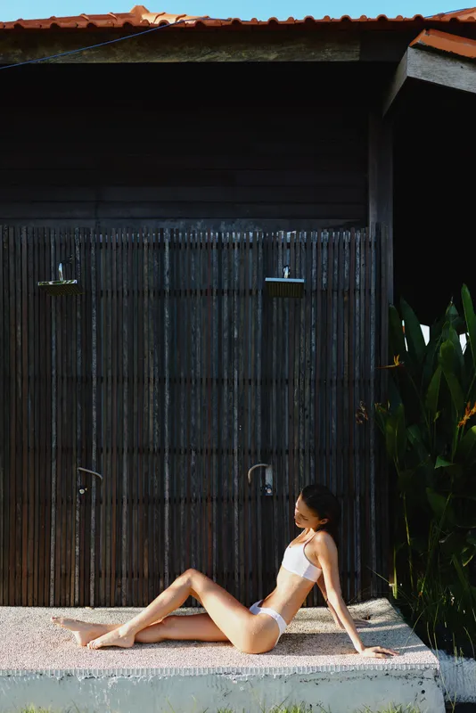 Young woman in white bikini sitting outdoors near wooden wall with outdoor showers, relaxed pose, sunny day, tropical plants, natural lifestyle, summer vacation concept