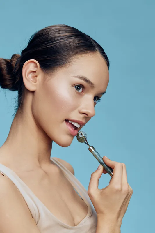 Young woman using a jade roller on her face, featuring a light blue background, promoting skincare, beauty routine, and healthy skin aesthetics