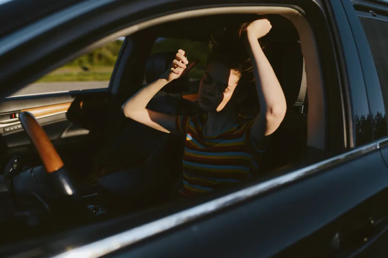 A woman sits in a car with her hands raised, captured in retro film tones. A candid lifestyle moment shows freedom, excitement, and bold style behind the wheel in warm light.