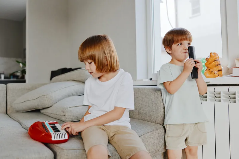 Two playful boys, one playing a toy keyboard, the other holding a microphone. Their joyful expressions highlight a fun moment at home, surrounded by cozy decor.