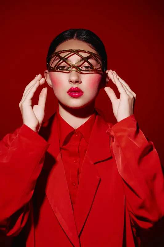 Young woman in red oversized blazer and shirt poses with lattice face accessory against a red background. Creative beauty and fashion concept with dramatic makeup and styling.