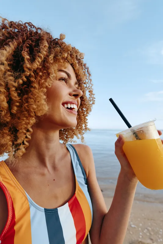 A joyful woman with curly hair drinks juice on the beach under a bright sky, wearing a colorful swimsuit, conveying carefree summer outdoor leisure.
