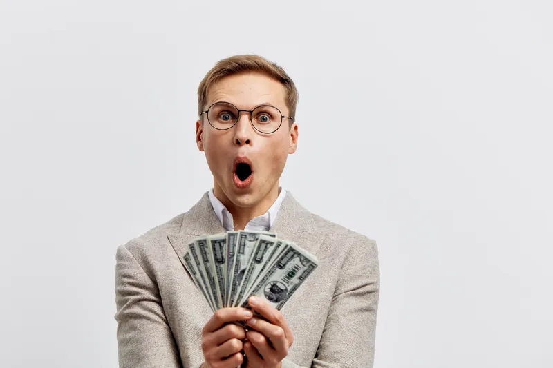 surprised young caucasian man hold us dollar bills wear beige jacket glasses isolated on gray background excited expression finance concept