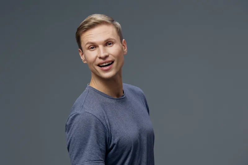 Cheerful young man with short blond hair wearing a casual blue sweater, smiling happily at camera, isolated on plain gray background, studio portrait. People lifestyle concept
