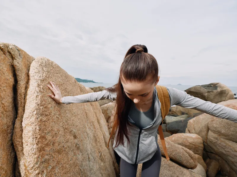 A young woman exploring rocky terrain at the beach with a focused expression, dressed in sporty attire, highlighting adventure and outdoor activity
