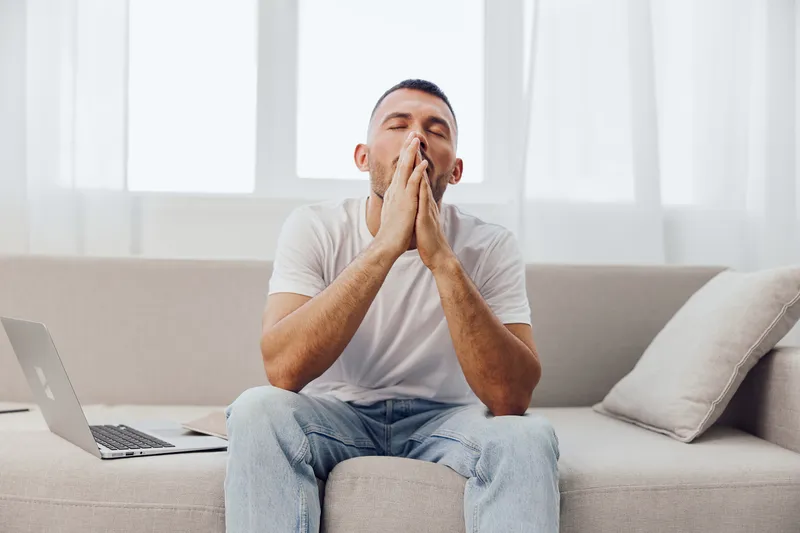 Tired man sitting on a sofa, expressing AI anxiety and emotional vulnerability in a casual home environment. Context reflects modern work-life blur and stress.