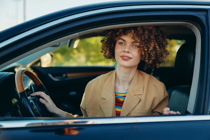 Smiling woman with curly hair sits in a vintage car, hands on the wheel. Retro film color tones, casual outfit, warm mood, everyday travel, self expression, cinematic vibe.