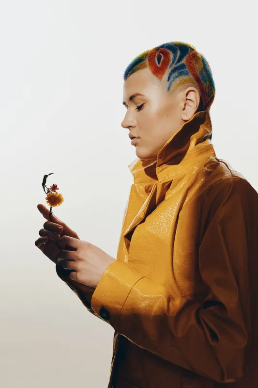 woman with colorful painted hair wearing a yellow jacket, holding a small flower, artistic style, side profile, minimalistic background, contemporary fashion