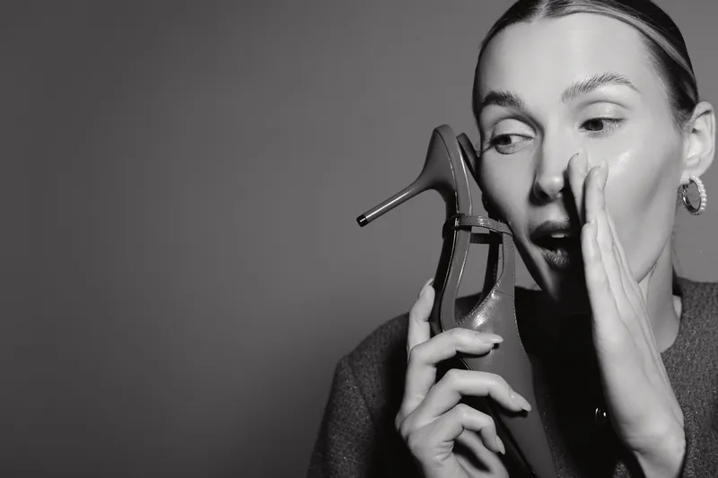 A fair skinned white woman with light hair holds a high heel shoe near her face and whispers with a surprised expression on a plain dark background.