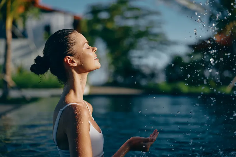 A young woman in a white swimsuit basks by a sunlit pool, her face raised to the spray as droplets fall, capturing a moment of calm and refreshment.