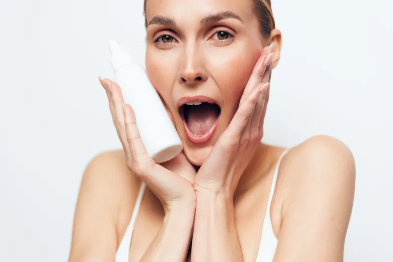Woman skincare face beauty surprised expression with cream, closeup on cheeks, young adult shoulders, studio white background, natural daylight, smooth skin, self care routine