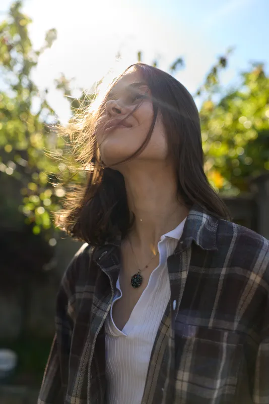 Casual woman outdoors, relaxed lifestyle moment, wind in hair, plaid shirt over white tee, natural sunlight, soft colors, candid portrait amid greenery, warm breeze, everyday fashion vibe