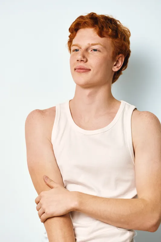 Young man with red hair and freckles wearing white tank top standing with crossed arms and looking away on white background in studio portrait.