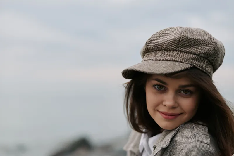 Young woman wearing a grey tweed newsboy cap and jacket smiles softly, looking at camera with blurred outdoor background. Concept of autumn fashion, casual style, and natural beauty.