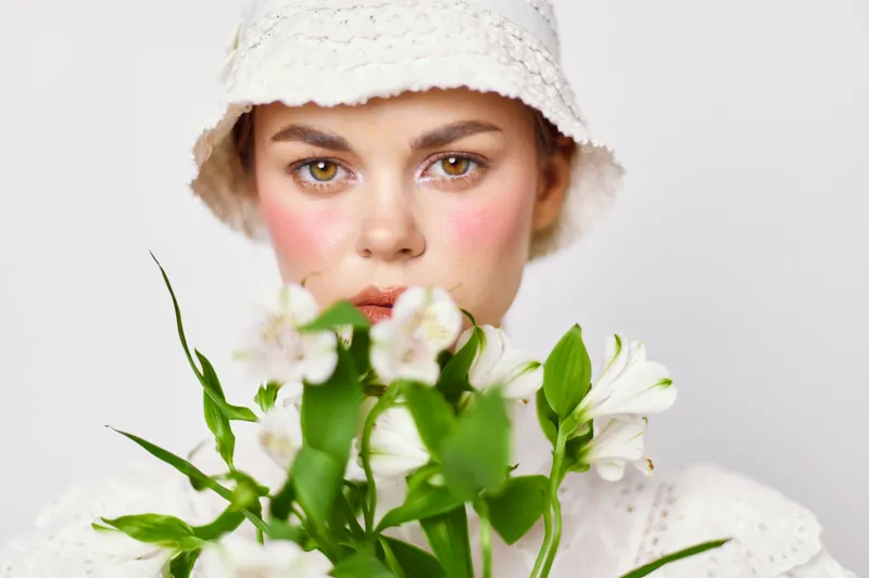 Young woman in white attire holding flowers, showcasing a serene expression against a light background, perfect for beauty and wellness themes