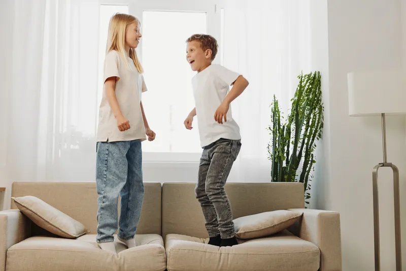 Two happy children jumping on beige sofa in bright living room with cushions and tall green plant near window. Concept of joy, playfulness, and family time indoors.
