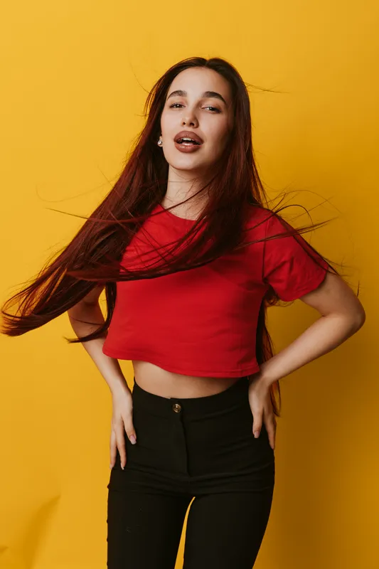 Young woman with long hair posing in red crop top and black pants against yellow background. Fashion, casual style and confident expression in studio portrait with motion in hair.