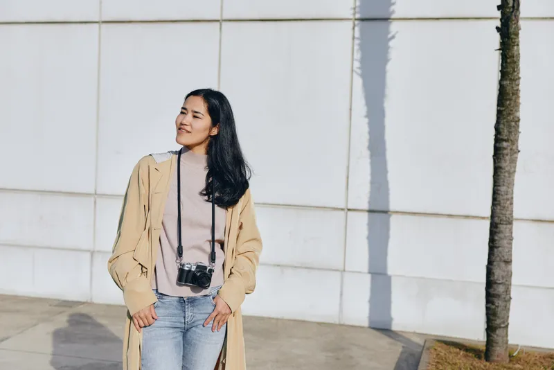 Female model in a beige coat and denim jeans stands beside a white wall with a camera, showcasing outdoor fashion, casual styling, and street photography in a bright daytime setting