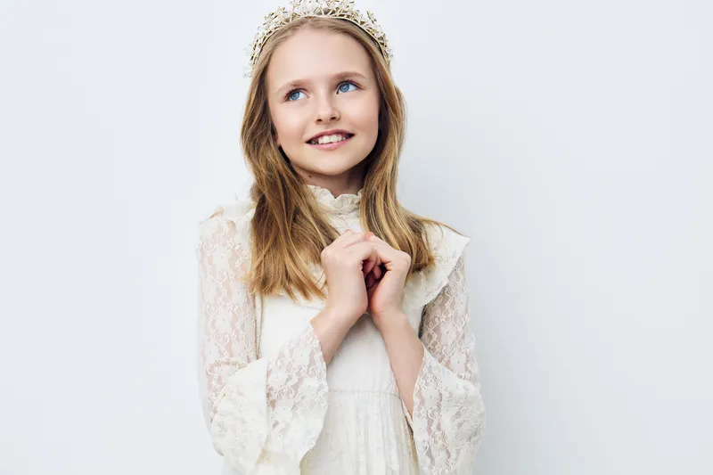 Young girl with a delicate crown, smiling in elegant white dress against a light pastel background Perfect for themes of innocence and childhood