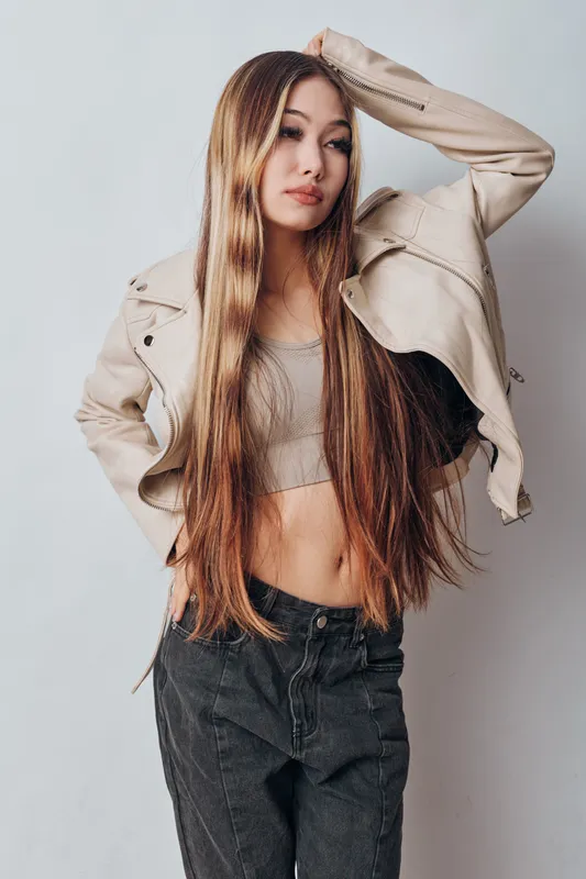 Young woman with long hair wearing beige jacket and crop top posing with hand on head against plain background. Fashion casual style and confident expression in studio.