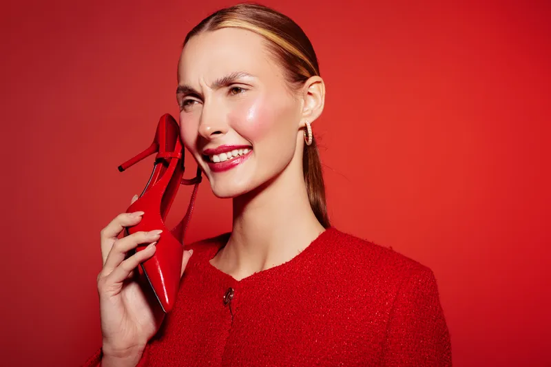 Smiling woman in red jacket holds red high heel shoe near face against red background. Concept of fashion, style, beauty, and creative portrait with vibrant color contrast and joyful mood.