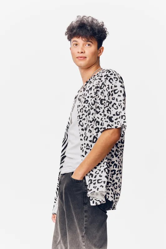 young man wearing leopard print shirt and casual pants standing relaxed with hands in pockets, short curly hair, neutral background, modern style outfit
