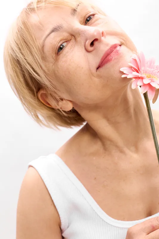Smiling blonde woman with short hair holding a pink flower in a beauty portrait, skincare and wellness concept, bright spa mood, soft studio light on white background.