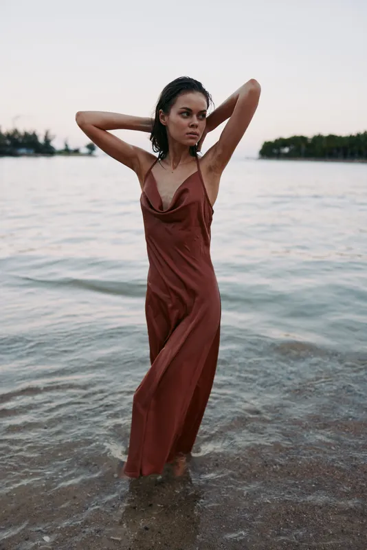 A fashion model stands waist deep in clear shallow water wearing a flowing rust dress, striking a confident pose with hands in hair, highlighting grace and movement.