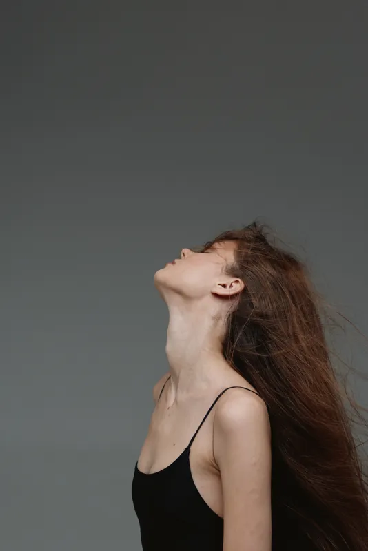 Young woman with long hair posing against grey background in black top, artistic and elegant portrait with soft lighting.