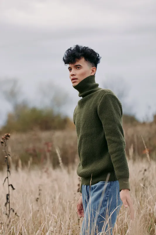 A young man stands in a grass field wearing a dark green knit turtleneck, exuding authenticity and quiet confidence in a natural portrait with soft light and minimalist backdrop.